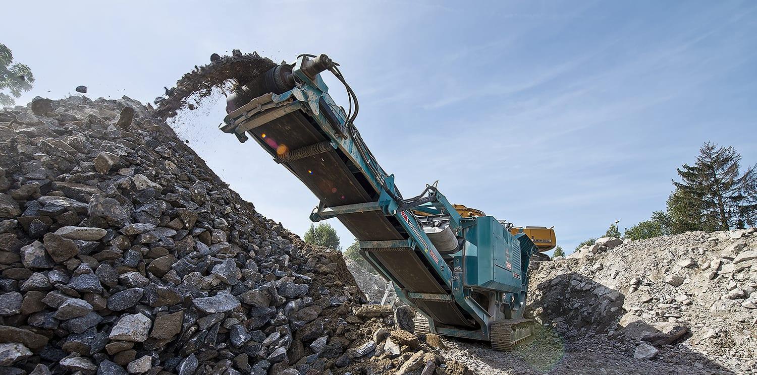 A tracked mobile crusher discharging crushed rock onto a stockpile in daylight.