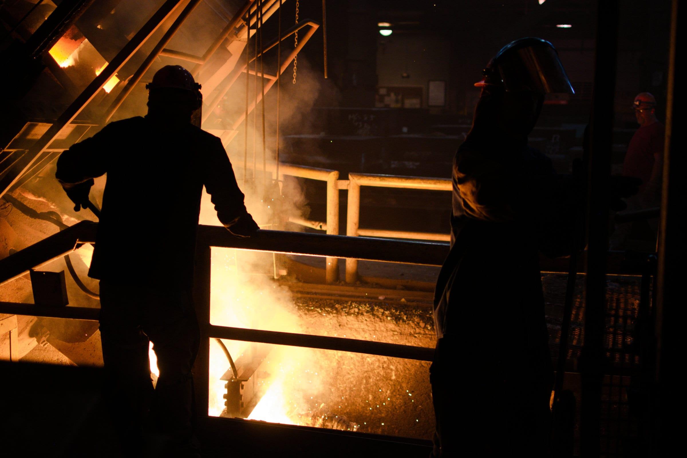 Two foundry workers in helmets and face shields silhouetted against the light of a pour.