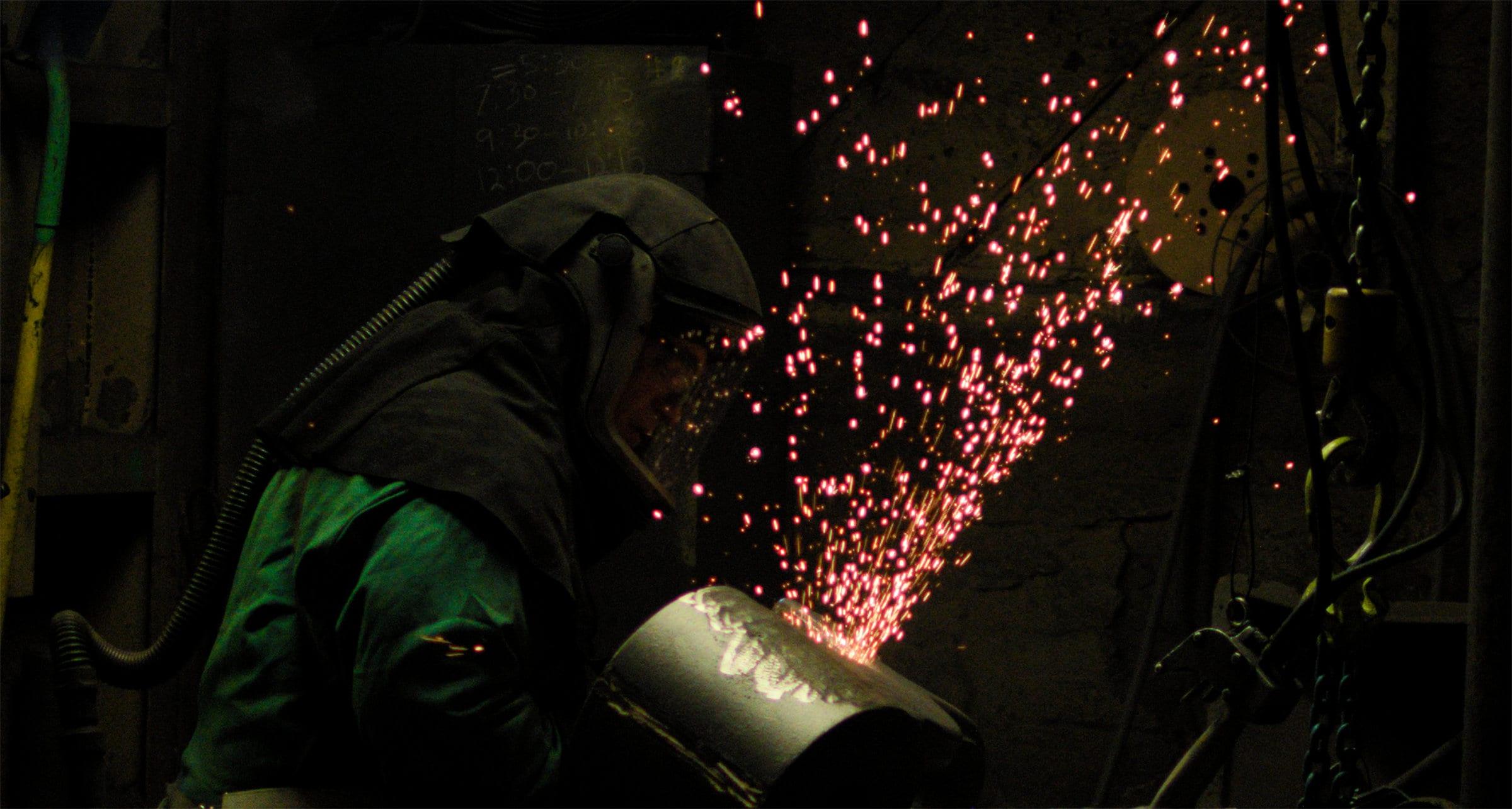 A worker in a hood gouging a casting, sparks arcing away across a dark bay.