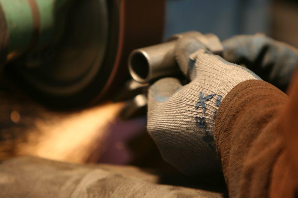 Gloved hands holding a small casting against a belt sander, sparks running off the contact point.