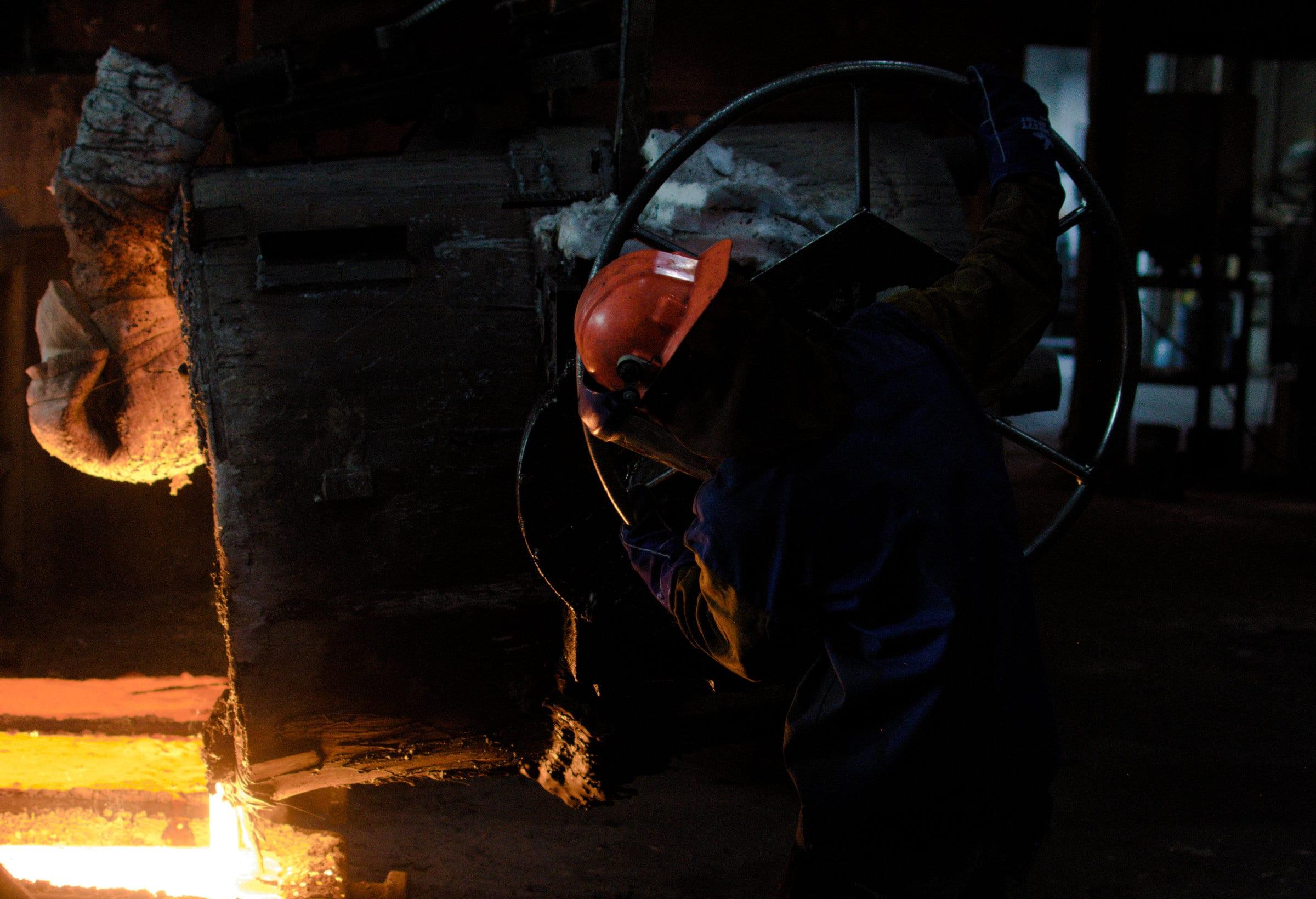 A worker leaning into the handwheel to tilt a ladle, metal running from the lip into the mold below.