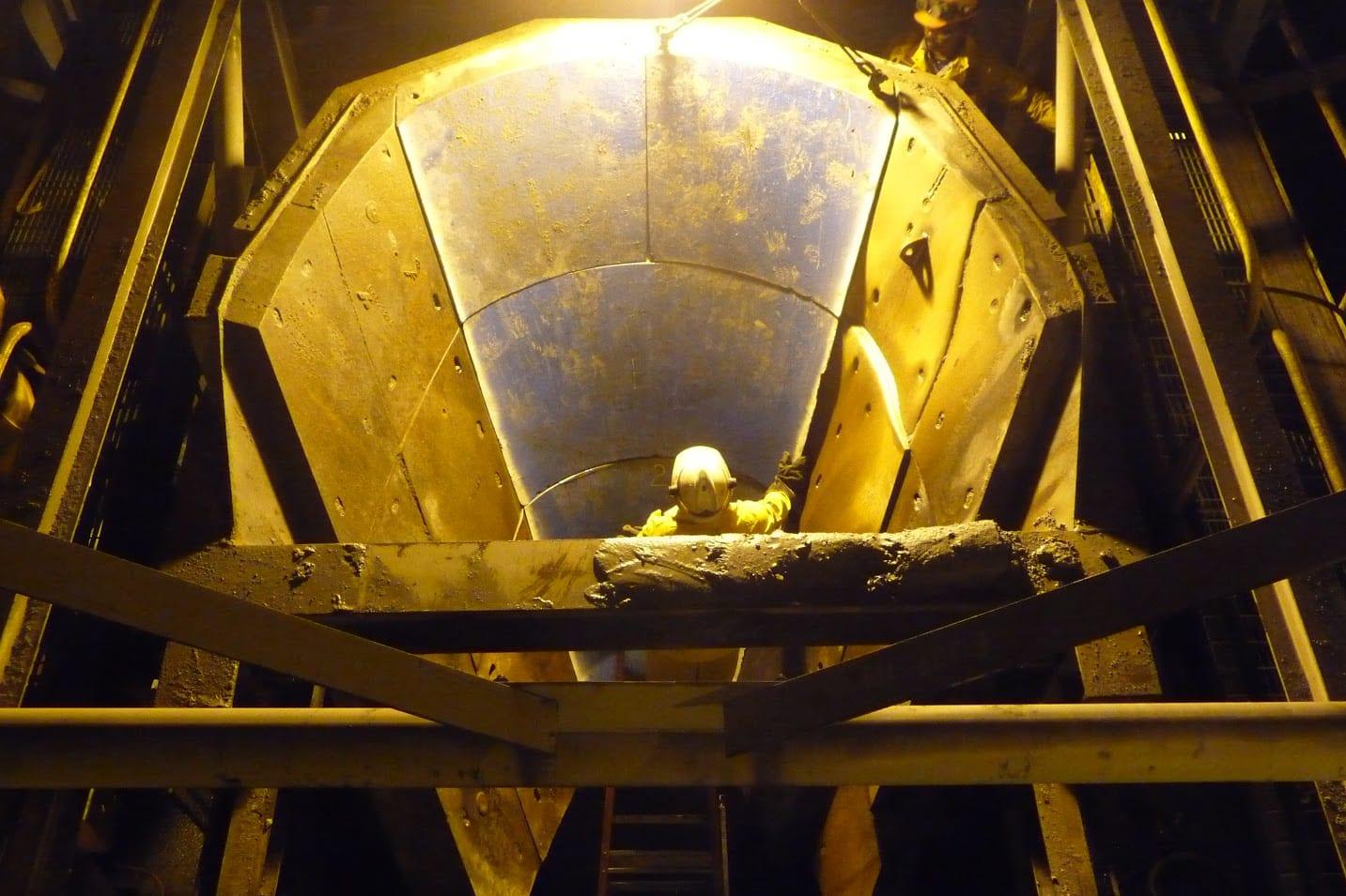 A worker inside a large ore transfer chute under sodium light, the walls lined with bolted wear segments.