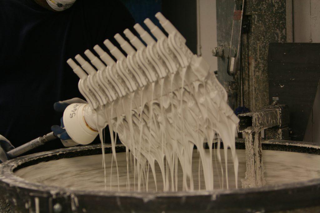 A wax pattern cluster lifted out of the ceramic slurry, coating running off it in strands.