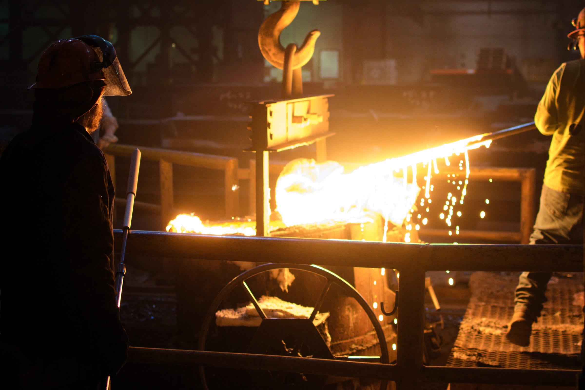 A white-hot stream of steel running from the furnace into a ladle, two workers watching from behind the rail.